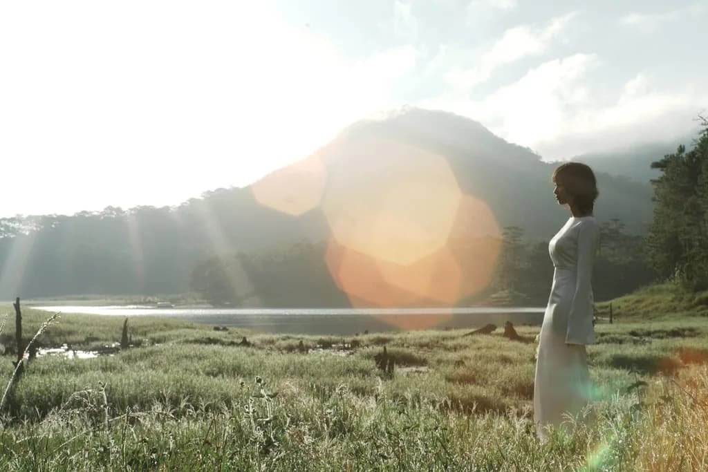 Hoàng Anh in a white áo dài, standing in a sunlit field by a lake