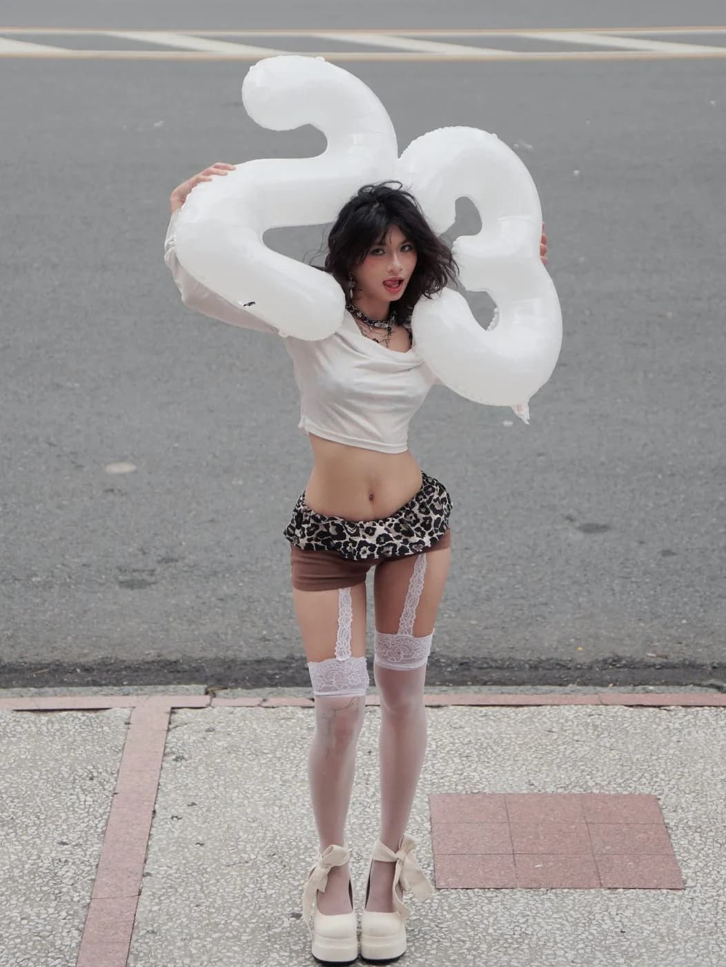 Hoàng Anh holding silver number balloons, in a white crop top, leopard mini, and platform heels