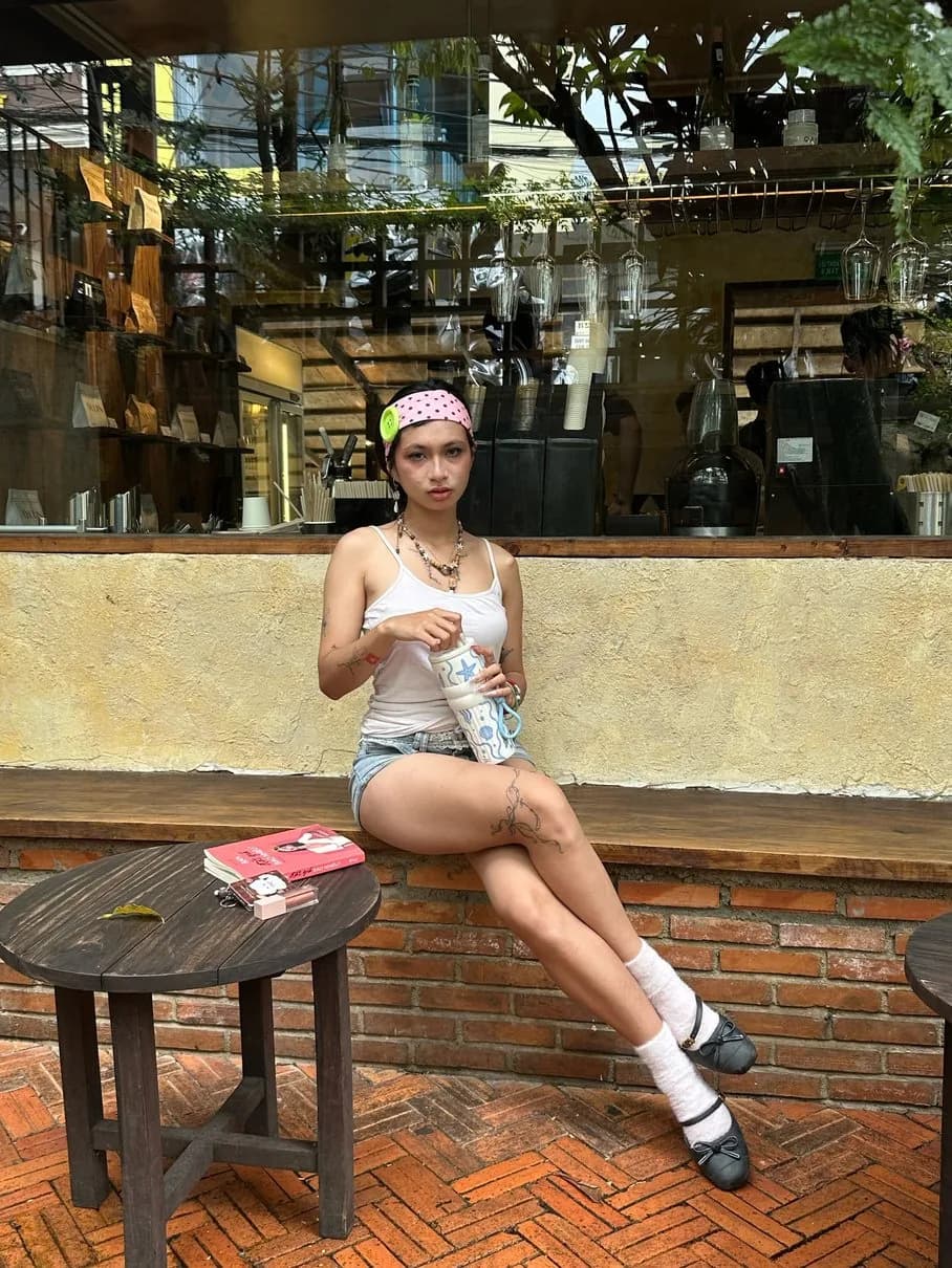 Hoàng Anh seated outside a café with a patterned cup, denim shorts, and a pink headband