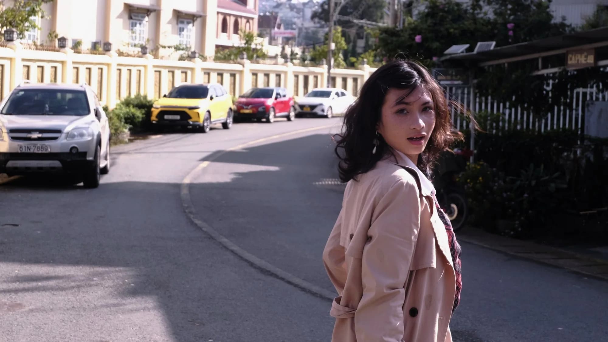 Hoàng Anh looking back on a curved street in Đà Lạt, beige trench over her shoulder