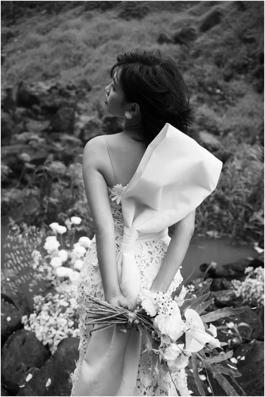 Hoàng Anh in a white lace dress, holding a bouquet beside dark rock and flowers