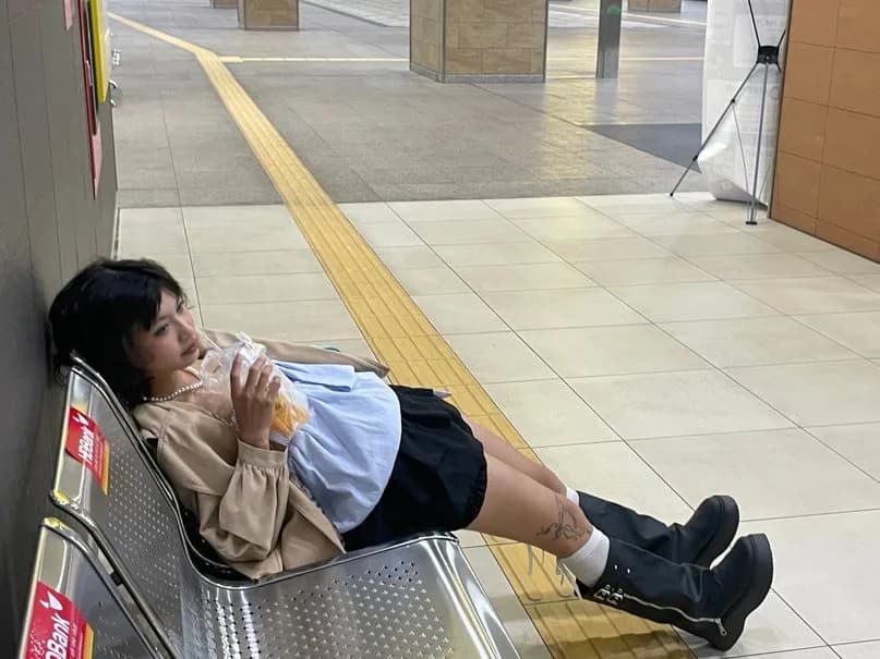 Hoàng Anh reclining across metal seats in a station, holding a drink