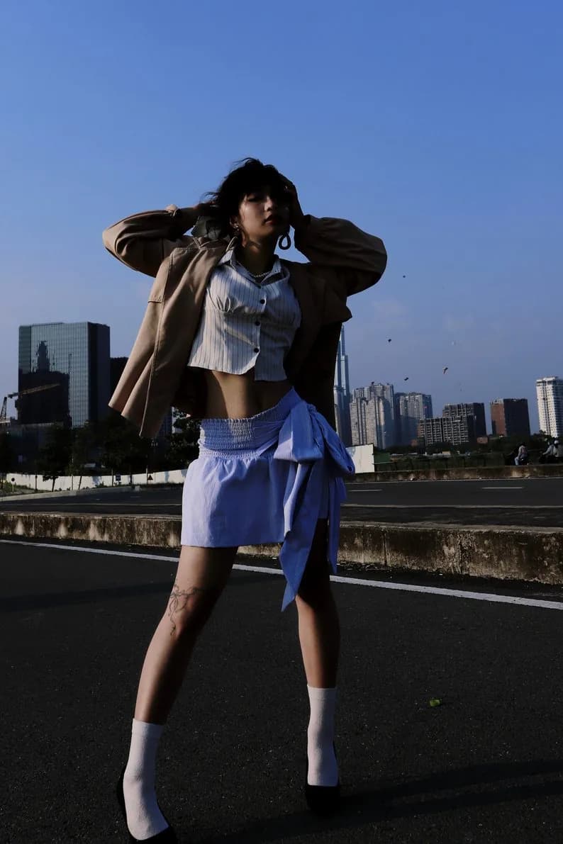 Hoàng Anh on a rooftop in a cropped shirt, blue skirt, and beige jacket, city skyline behind her