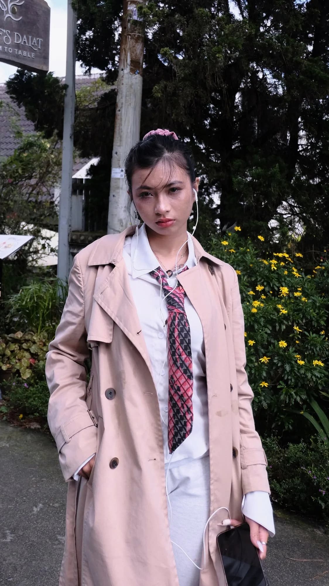 Hoàng Anh in a beige trench, white shirt, and patterned tie, earphones in, standing in Đà Lạt